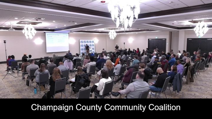 Wide shot from the back of the room of the audience, the presenter, and the presentation screen. 